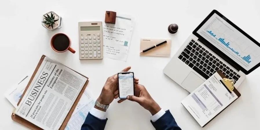 Company Setup Dubai - A person at a desk with a business newspaper, smartphone, laptop, coffee mug, and office supplies.