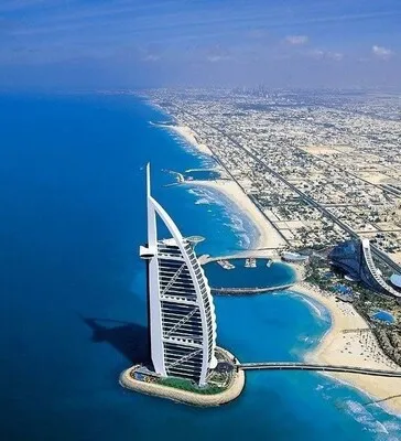 business set up companies in dubai  - Aerial view of a sail-shaped skyscraper by the coastline with a clear blue sea and city in the background.